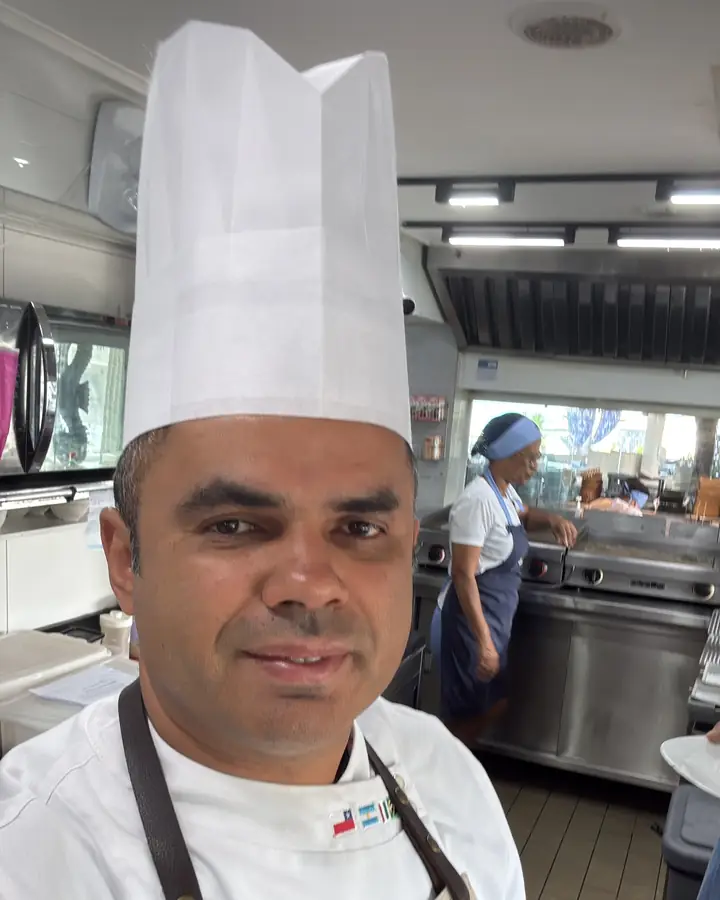 Joel Pinheiro de dólmã e toque blanche na cozinha, com a equipe em produção ao fundo